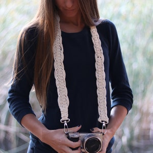 Puede incluir: Una mujer con una camisa azul oscuro y pantalones negros sostiene una cámara plateada con una correa de cámara blanca. La correa de la cámara está hecha de cuerda trenzada.