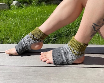 May include: A pair of knit yoga socks featuring an open-toe and open-heel design. The socks are constructed with a ribbed cuff in olive green, followed by a band of white, and a dark gray section covering the mid-foot and arch area.