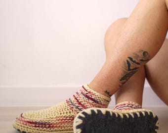 Puede incluir: Un par de zapatillas de estar por casa a la altura del tobillo con un tejido de punto color crema y rayas horizontales en rojo y azul. Las zapatillas tienen una suela de fieltro gris oscuro unida con puntadas de festón color crema.