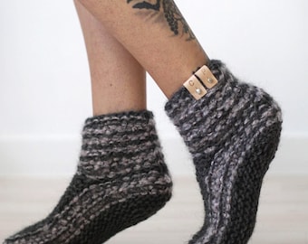 May include: A pair of ankle-length knitted socks in a dark charcoal and light gray variegated yarn. The socks feature a thick, textured knit pattern and include small, rectangular tan leather tags attached to the cuffs with silver metal rivets.