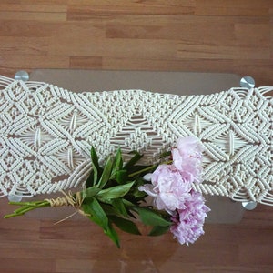 May include: A white macrame table runner with a geometric pattern, featuring a bouquet of pink peonies on a glass table.