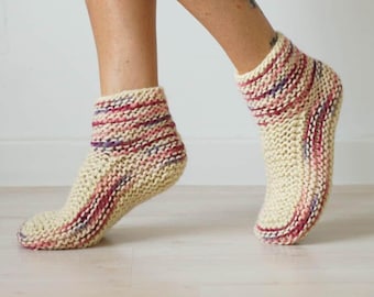 May include: A pair of ankle-length hand-knitted socks made from thick cream-colored yarn. The socks feature horizontal stripes of variegated pink, purple, and red yarn integrated into the knit pattern, creating a textured and chunky appearance.
