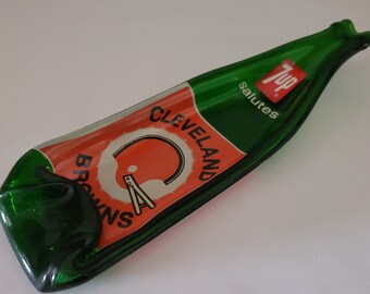 May include: A green glass 7 Up bottle shaped dish with a red, white, and orange Cleveland Browns logo. The 7 Up logo is red and white and says "Salutes".