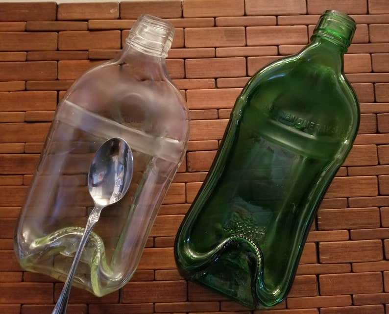 May include: Two glass bottle spoon rests, one clear and one green. The clear bottle rest has a silver spoon inside. The green bottle rest has a label that says "Sandqvist" on it.