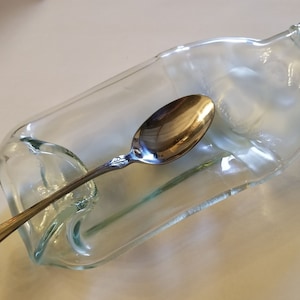 May include: A clear glass bottle with the top cut off and a spoon resting inside. The bottle is shaped like a bowl and is used as a spoon rest.