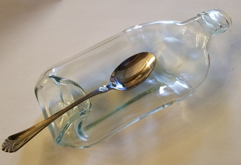 May include: A clear glass bottle with the top cut off and a spoon resting inside. The bottle is shaped like a bowl and is used as a spoon rest.