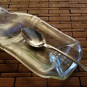 May include: A clear glass bottle, shaped like a spoon rest, with a silver spoon resting inside.