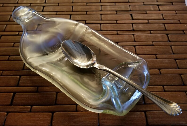 May include: A clear glass bottle, shaped like a spoon rest, with a silver spoon resting inside.