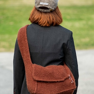 May include: A fuzzy brown knit crossbody bag with a flap closure and a silver-toned safety pin accent. The bag is worn over a black blazer, paired with a distressed grey baseball cap featuring the word BALMAIN embroidered in white on the back.