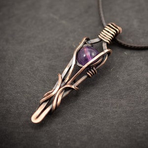 May include: A copper wire wrapped pendant with an amethyst gemstone. The pendant is attached to a brown leather cord. The pendant is a unique and handmade piece of jewelry.