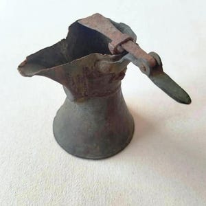 Puede incluir: Una jarra de metal antigua con pico y asa. La jarra tiene una parte superior acampanada y una base redondeada. El metal tiene un aspecto desgastado y envejecido, con zonas de óxido y decoloración. El asa está sujeta con una bisagra.