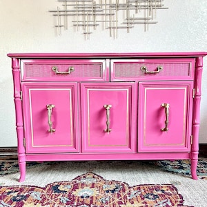 May include: A bright pink cabinet with gold accents. The cabinet has two drawers with woven fronts and brass handles. The doors have gold trim and ornate brass handles. The cabinet is set on curved legs and has a bamboo-style frame.