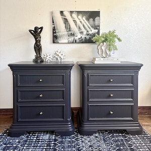 May include: Two dark navy blue nightstands, each featuring three drawers with vertical ribbed detailing and small brass-toned knobs. The nightstands have a rectangular top, a sturdy base, and a smooth matte finish, providing storage space.