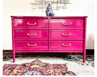 May include: A bright pink dresser with six drawers and gold handles. The dresser is made of faux bamboo and has a glossy finish. It is sitting on a rug with a floral pattern.