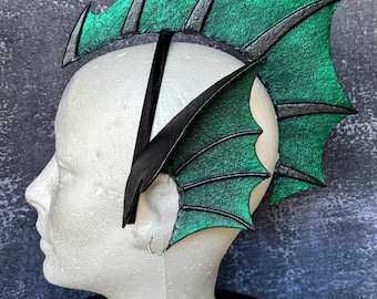 Può includere: Un copricapo caratterizzato da una grande struttura a pinna arcuata in verde metallizzato con bordi neri, fissata a un cerchietto nero. Il set include pinne laterali per le orecchie abbinate. Il testo recita: Headfin with Mermaid ear fins. This photo shows the color closest to how it looks in person.