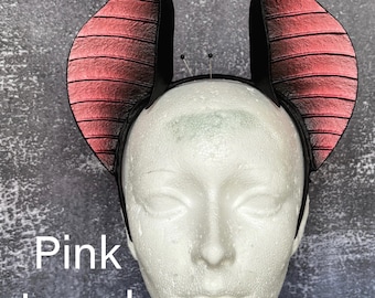 May include: A headband featuring two large, upright bat-like ears with a textured, pink-toned gradient finish that transitions to black edges. The ears have horizontal ridges and are mounted on a thin black band. The text Pink toned is visible.