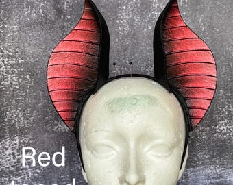 May include: A headband featuring two large, curved, horn-like structures with a textured red and black gradient finish. The horns are mounted on a thin black band. The text "Red toned" is visible on the left side of the image.