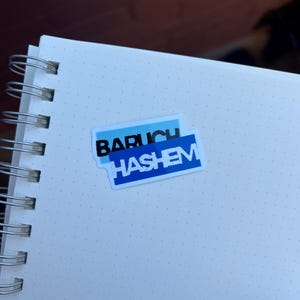 Può includere: Un quaderno bianco a spirale con un adesivo blu e bianco che recita "BARUCH HASHEM". L'adesivo è rettangolare con sfondo blu e testo bianco. Il quaderno ha un motivo a griglia puntinata.
