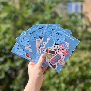 May include: A hand holding several blue sticker packs. Each pack has a round sticker that says "OOPS!" and contains assorted stickers with minor cosmetic defects. The stickers feature various designs and text.