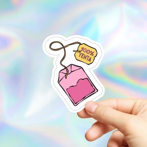 May include: A pink tea bag sticker with the text "100% YENTA" on a yellow tag. The tea bag is filled with a darker shade of pink and has a string attached. The sticker is held by a hand against a holographic background.