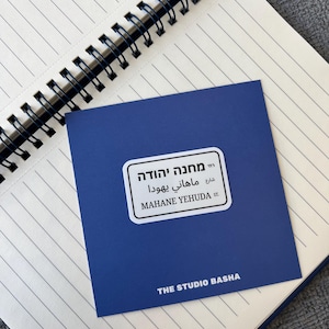 Peut inclure: Une carte bleue avec le texte "Mahane Yehuda St." en anglais et en hébreu, posée sur un carnet ligné. La carte porte également le texte "THE STUDIO BASHA". Le carnet a une reliure à spirale noire.