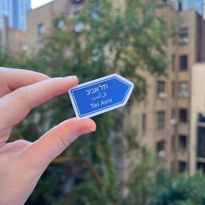 Peut inclure: Un autocollant bleu et blanc en forme de panneau routier, avec le texte "Tel Aviv" en hébreu et en anglais. L'autocollant est tenu devant un arrière-plan flou de bâtiments et d'arbres.