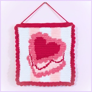 May include: A handmade crochet wall hanging featuring a large red heart design. The heart is layered with shades of pink and red, set against a striped background of white and light pink. The edges are trimmed with a scalloped red border.