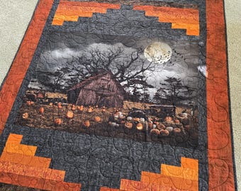 May include: A Halloween-themed quilt with a patchwork design. The center panel features a spooky scene with a farmhouse, trees, and a full moon. The border is made of orange and black fabric with a quilted pattern.