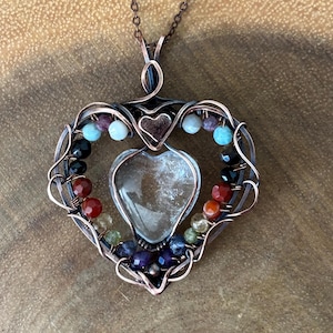 May include: A heart-shaped pendant necklace crafted from dark copper wire, featuring a central clear heart-shaped stone. The wire frame is adorned with small, multi-colored faceted beads in shades of blue, black, red, yellow, and purple.