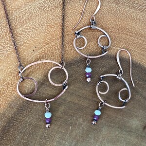 May include: A copper-toned necklace and matching pair of earrings featuring the Cancer zodiac symbol. Each piece is crafted from wire shaped into a circle with two smaller internal loops, accented with a light blue, dark pink, and metallic bead.