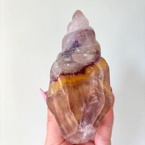 May include: A translucent, carved crystal sculpture in the shape of a spiral seashell. The crystal features a blend of colors, including lavender, yellow, and amber, with darker purple and brown veining. The sculpture is held in a hand.