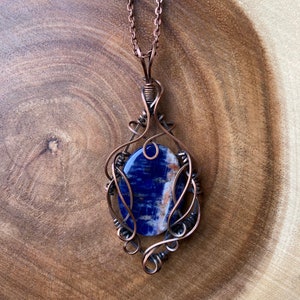 May include: A copper wire wrapped pendant featuring a blue and white gemstone. The pendant is attached to a copper chain.