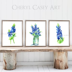 Puede incluir: Tres acuarelas enmarcadas de flores de lupino azul. Las pinturas de la izquierda y la derecha presentan una sola flor de lupin azul, mientras que la pintura del centro muestra un ramo de lupinos azules en un frasco de vidrio transparente.