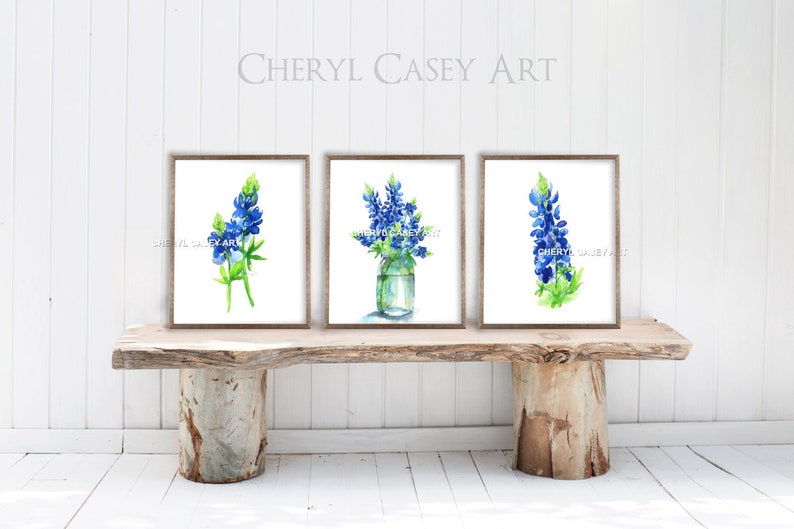 Puede incluir: Tres acuarelas enmarcadas de flores de lupino azul. Las pinturas de la izquierda y la derecha presentan una sola flor de lupin azul, mientras que la pintura del centro muestra un ramo de lupinos azules en un frasco de vidrio transparente.
