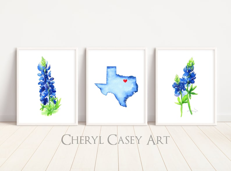 May include: Three watercolor paintings in white frames. The left and right paintings feature bluebonnet flowers. The center painting features a blue watercolor outline of the state of Texas with a red heart in the center.