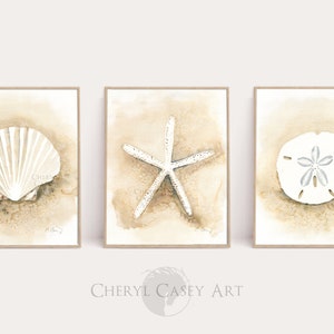 Puede incluir: Conjunto de tres acuarelas que representan una concha, una estrella de mar y un dólar de arena sobre un fondo beige. Las pinturas están enmarcadas en un marco de madera claro.
