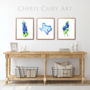 May include: Three framed watercolor paintings on a white wall above a wooden console table. The paintings feature a bluebonnet flower, the state of Texas with a red heart in the center, and another bluebonnet flower. The console table has two black wire baskets on the bottom shelf. The text "welcoming home" is on a pillow in one of the baskets.