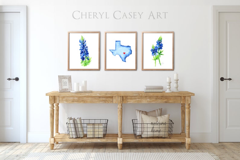 May include: Three framed watercolor paintings on a white wall above a wooden console table. The paintings feature a bluebonnet flower, the state of Texas with a red heart in the center, and another bluebonnet flower. The console table has two black wire baskets on the bottom shelf. The text "welcoming home" is on a pillow in one of the baskets.