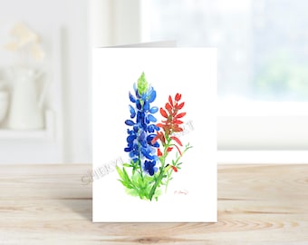 Peut inclure: Une carte de vœux à l'aquarelle avec une illustration vibrante de lupins bleus et de fleurs sauvages rouges. L'œuvre d'art présente les fleurs avec un feuillage vert sur fond blanc. La carte est posée sur une surface en bois.