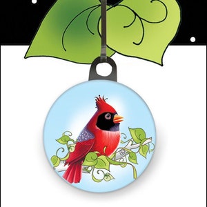 May include: A red cardinal bird with a black head and a yellow beak perched on a branch with green leaves. The bird is on a round, blue background with a black zipper pull attached.
