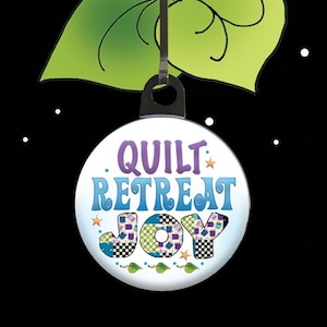 May include: A white round zipper pull charm with the text "QUILT RETREAT JOY" in colorful letters. The letters are decorated with a patchwork pattern. The charm is attached to a black metal ring.