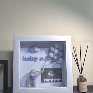 Puede incluir: Un marco blanco con un elefante de ganchillo sosteniendo globos azules y blancos. El texto "baby a" está en ganchillo azul. También se incluye una imagen de ultrasonido.