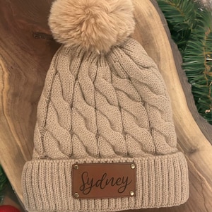 May include: A beige knitted beanie with a brown faux fur pom pom and a brown leather patch with the name "Sydney" on it.