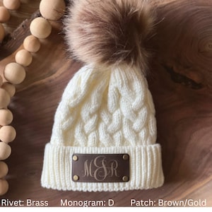 May include: White cable knit beanie with a brown faux fur pom pom and a brown leather patch with the monogram "MSR" in gold lettering.
