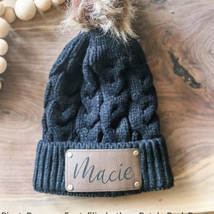 May include: Black knit beanie with a faux fur pom pom and a brown leather patch that says "Macie" in a cursive font. The patch has brass rivets.