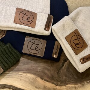May include: Four beige knit beanies with brown leather patches. Each patch has a circle with the letters "te" inside and a name below. The names are Tanner, Tanner, Bird, and a fourth name that is unclear.