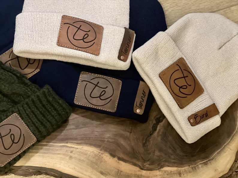 May include: Four beige knit beanies with brown leather patches. Each patch has a circle with the letters "te" inside and a name below. The names are Tanner, Tanner, Bird, and a fourth name that is unclear.