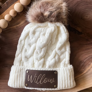 Peut inclure: Bonnet en tricot blanc à câble avec un pompon en fausse fourrure marron et un patch en cuir marron avec le nom "Willow" en lettres dorées.