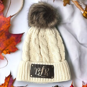 May include: Cream colored knitted beanie with a brown faux fur pom pom. The beanie has a brown leather patch with gold lettering that reads "B.L.R."