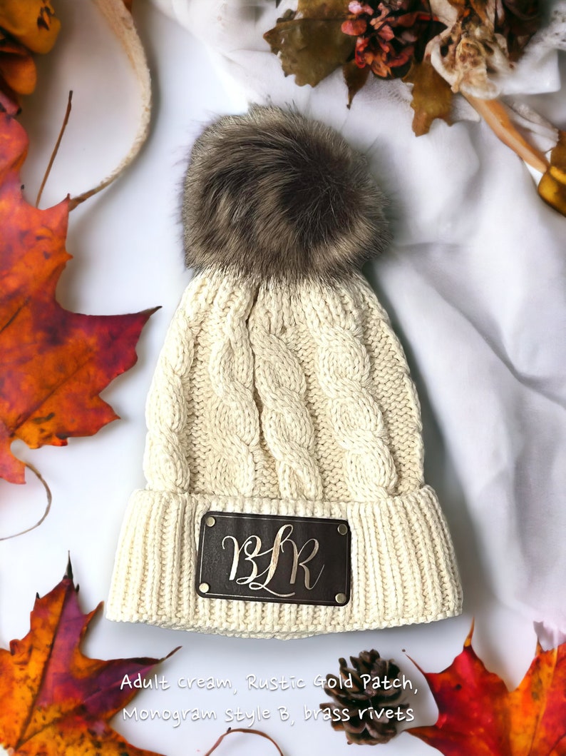 May include: Cream colored knitted beanie with a brown faux fur pom pom. The beanie has a brown leather patch with gold lettering that reads "B.L.R."